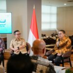 Panel discussion with three men seated in chairs, Indonesian flag between them and a screen showing event text on the left.