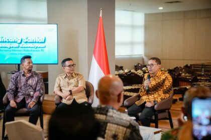 Panel discussion with three men seated in chairs, Indonesian flag between them and a screen showing event text on the left.