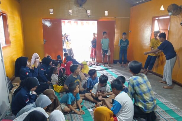 Group of children seated on mats in a yellow-walled room while several teens perform at the doorway area to the right.