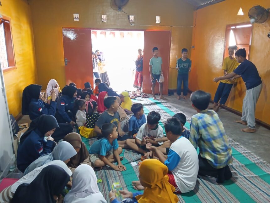 Group of children seated on mats in a yellow-walled room while several teens perform at the doorway area to the right.