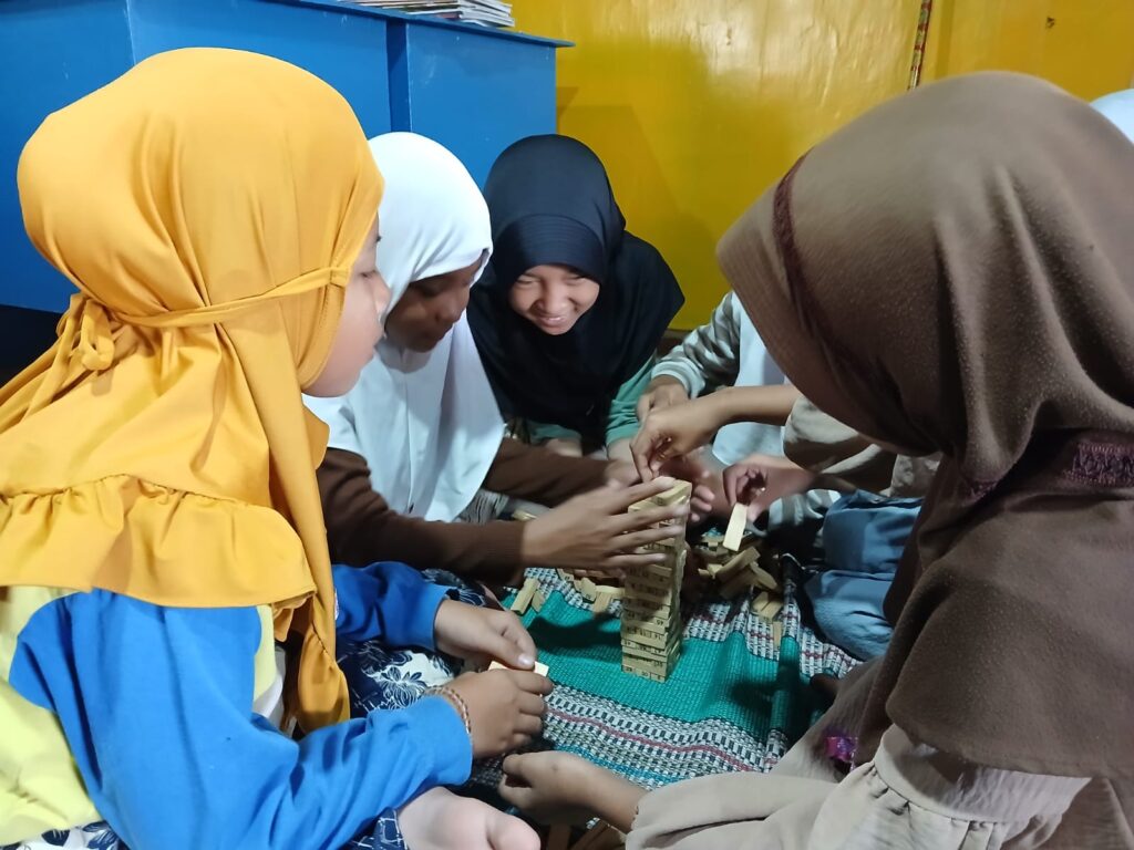 Group of girls in colorful hijabs building a wooden block tower on a green rug in a bright room.