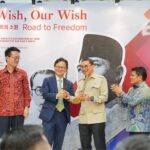 Four men on a stage at an exhibition, exchanging a ceremonial token while others applaud, with a large banner that reads 'My Wish, Our Wish' in the background.