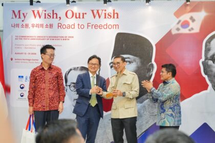 Four men on a stage at an exhibition, exchanging a ceremonial token while others applaud, with a large banner that reads 'My Wish, Our Wish' in the background.