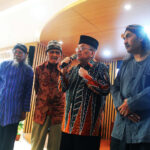 Four older men in traditional clothing sing or speak on a stage in a bright room, one holding a microphone.
