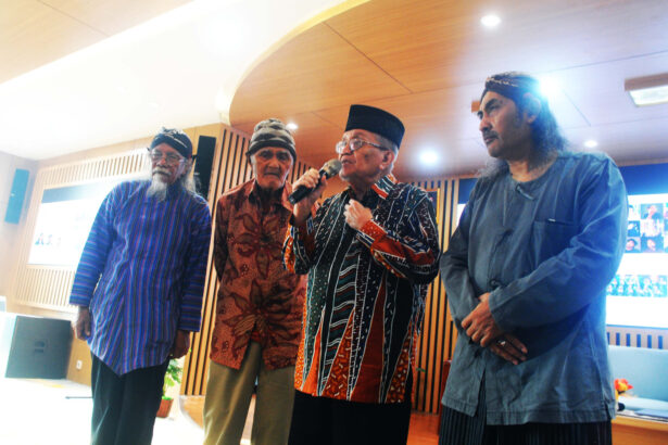 Four older men in traditional clothing sing or speak on a stage in a bright room, one holding a microphone.