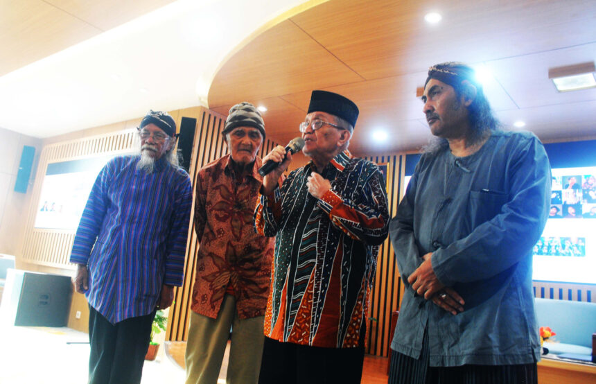 Four older men in traditional clothing sing or speak on a stage in a bright room, one holding a microphone.