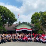 Large group of students and teachers posing for a photo in front of SDN Margorejo school, with an Indonesian flag and red-white bunting overhead.