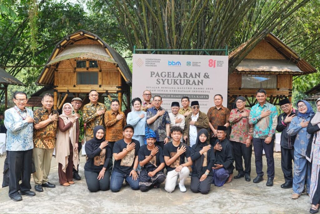 WhatsApp Image 2026-08-15 at 163646 - Mabur.co Group of diverse Indonesian people posing for a photo at an outdoor cultural event, standing in front of a banner that reads 'Pagelaran & Syukuran', with many placing a hand on the chest in a ceremonial gesture.