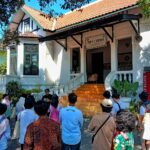 Group of students and visitors gathered outside a white colonial-style building with orange-tiled roof and steps leading to an open entrance.