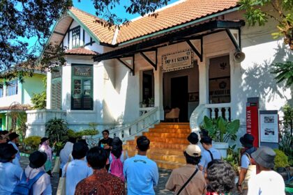 Group of students and visitors gathered outside a white colonial-style building with orange-tiled roof and steps leading to an open entrance.