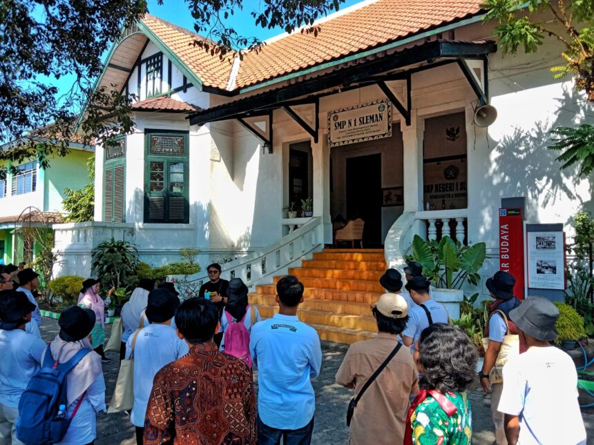 Group of students and visitors gathered outside a white colonial-style building with orange-tiled roof and steps leading to an open entrance.