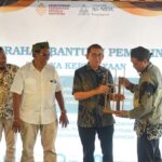Four men stand on a stage during a government aid handover ceremony; one man receives a wooden model from another, with a banner and logos in the background.