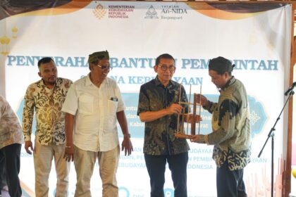 Four men stand on a stage during a government aid handover ceremony; one man receives a wooden model from another, with a banner and logos in the background.