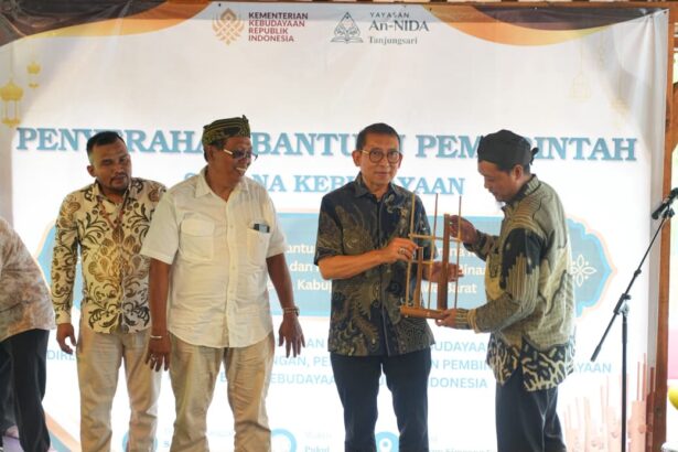 Four men stand on a stage during a government aid handover ceremony; one man receives a wooden model from another, with a banner and logos in the background.