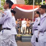 Group of cadets in white uniforms marching past a crowd under a red-and-white tent backdrop.