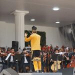 Conductor in a gold traditional outfit leads an orchestra on stage with string players and sheet music stands surrounding him.