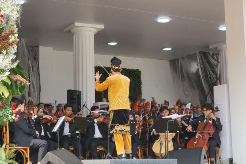 Conductor in a gold traditional outfit leads an orchestra on stage with string players and sheet music stands surrounding him.