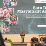 Speaker in batik at podium at an Indonesian conference; large banner reads 'Satu Data Masyarakat Adat' with collage images of indigenous communities.