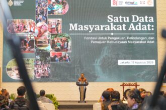 Speaker in batik at podium at an Indonesian conference; large banner reads 'Satu Data Masyarakat Adat' with collage images of indigenous communities.