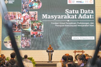 Speaker in batik at podium at an Indonesian conference; large banner reads 'Satu Data Masyarakat Adat' with collage images of indigenous communities.