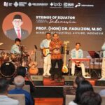 Stage scene of a formal musical event: a man in batik attire speaks at a microphone while a blue-jacketed band accompanies him; audience seated in front, backdrop shows event title 'Strings of Equator' and logos.