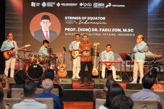 Stage scene of a formal musical event: a man in batik attire speaks at a microphone while a blue-jacketed band accompanies him; audience seated in front, backdrop shows event title 'Strings of Equator' and logos.