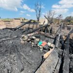Charred remains of a ruined room with blackened wooden beams and ash-covered floor, scattered books amid the rubble after a fire.