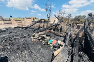 Charred remains of a ruined room with blackened wooden beams and ash-covered floor, scattered books amid the rubble after a fire.