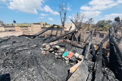 Charred remains of a ruined room with blackened wooden beams and ash-covered floor, scattered books amid the rubble after a fire.