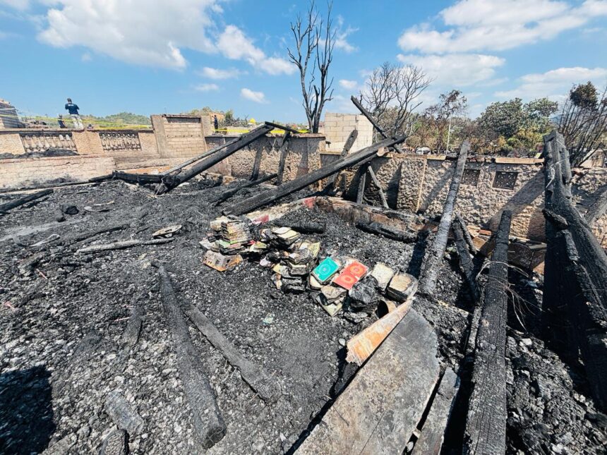 Charred remains of a ruined room with blackened wooden beams and ash-covered floor, scattered books amid the rubble after a fire.