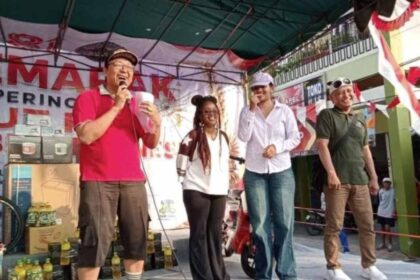Man in a bright pink shirt speaks into a microphone on a stage, with three others smiling beside him at an outdoor event. Bottles and boxes are on the ground behind them.