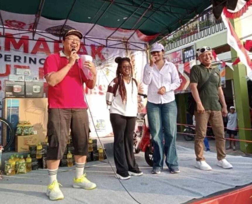 Man in a bright pink shirt speaks into a microphone on a stage, with three others smiling beside him at an outdoor event. Bottles and boxes are on the ground behind them.