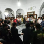 Large crowd of men and boys forming a circle in a white arched hall during a community gathering, center of attention unclear