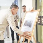 Man in a light shirt signs or places a note on a framed certificate on an easel as photographers document the moment.