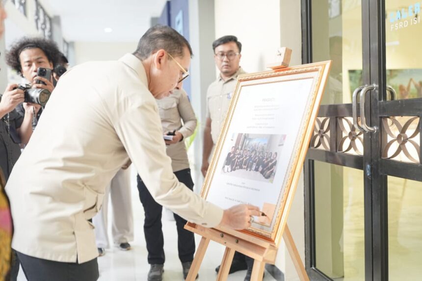 Man in a light shirt signs or places a note on a framed certificate on an easel as photographers document the moment.