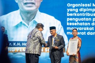 Two men on a stage shake hands as one receives a glass trophy; a woman stands nearby holding a framed certificate against a blue backdrop with text to the side.