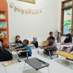 Teacher in a black hijab reads to a group of young students seated at small desks in a cozy classroom with bookshelves and bunting on the wall.
