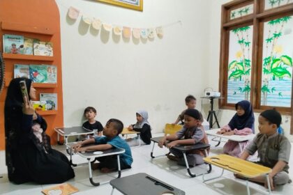 Teacher in a black hijab reads to a group of young students seated at small desks in a cozy classroom with bookshelves and bunting on the wall.