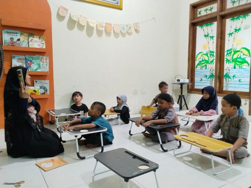 Teacher in a black hijab reads to a group of young students seated at small desks in a cozy classroom with bookshelves and bunting on the wall.
