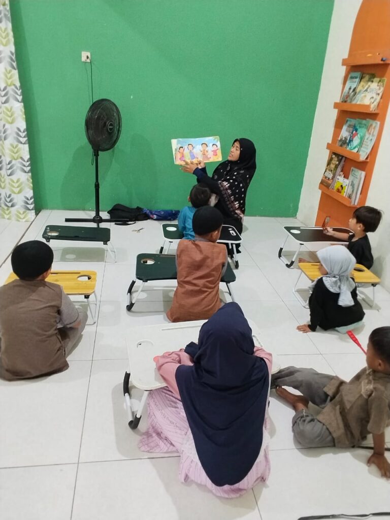 WhatsApp Image 2026-08-27 at 111444 - Mabur.co Teacher reads a picture book aloud to children seated on small desks in a bright classroom with a green wall behind them.