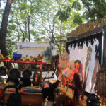 Outdoor cultural performance setup under trees, featuring a gold dragon prop atop a red frame with gongs and musicians nearby, and a large decorative backdrop behind them.