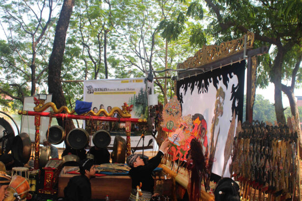 Outdoor cultural performance setup under trees, featuring a gold dragon prop atop a red frame with gongs and musicians nearby, and a large decorative backdrop behind them.