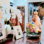 A man in a white shirt gazes at a memorial altar with framed photos, white orchids, and a fruit basket as a woman stands nearby in the background.