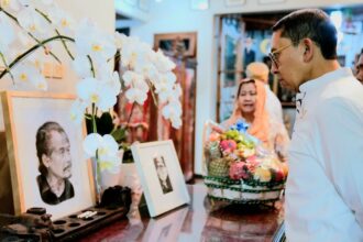 A man in a white shirt gazes at a memorial altar with framed photos, white orchids, and a fruit basket as a woman stands nearby in the background.