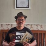 Smiling man wearing a hat and glasses sits on a wooden bench, holding three books in front of a framed certificate on the wall behind him.