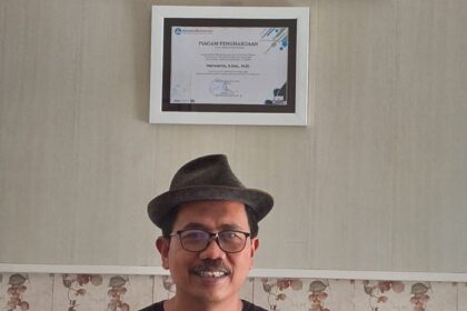 Smiling man wearing a hat and glasses sits on a wooden bench, holding three books in front of a framed certificate on the wall behind him.