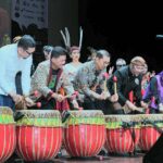 Group of diverse performers on stage playing large red and yellow drums with mallets during a cultural performance.