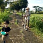 A young boy in a black shirt and tan shorts holds a turquoise bowl while walking down a rural path, an elderly man pushes a bicycle beside tall grass and trees in a sunny field.