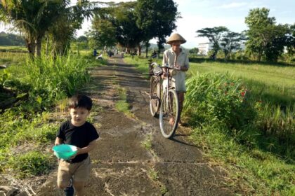 A young boy in a black shirt and tan shorts holds a turquoise bowl while walking down a rural path, an elderly man pushes a bicycle beside tall grass and trees in a sunny field.
