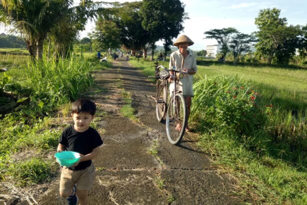 A young boy in a black shirt and tan shorts holds a turquoise bowl while walking down a rural path, an elderly man pushes a bicycle beside tall grass and trees in a sunny field.